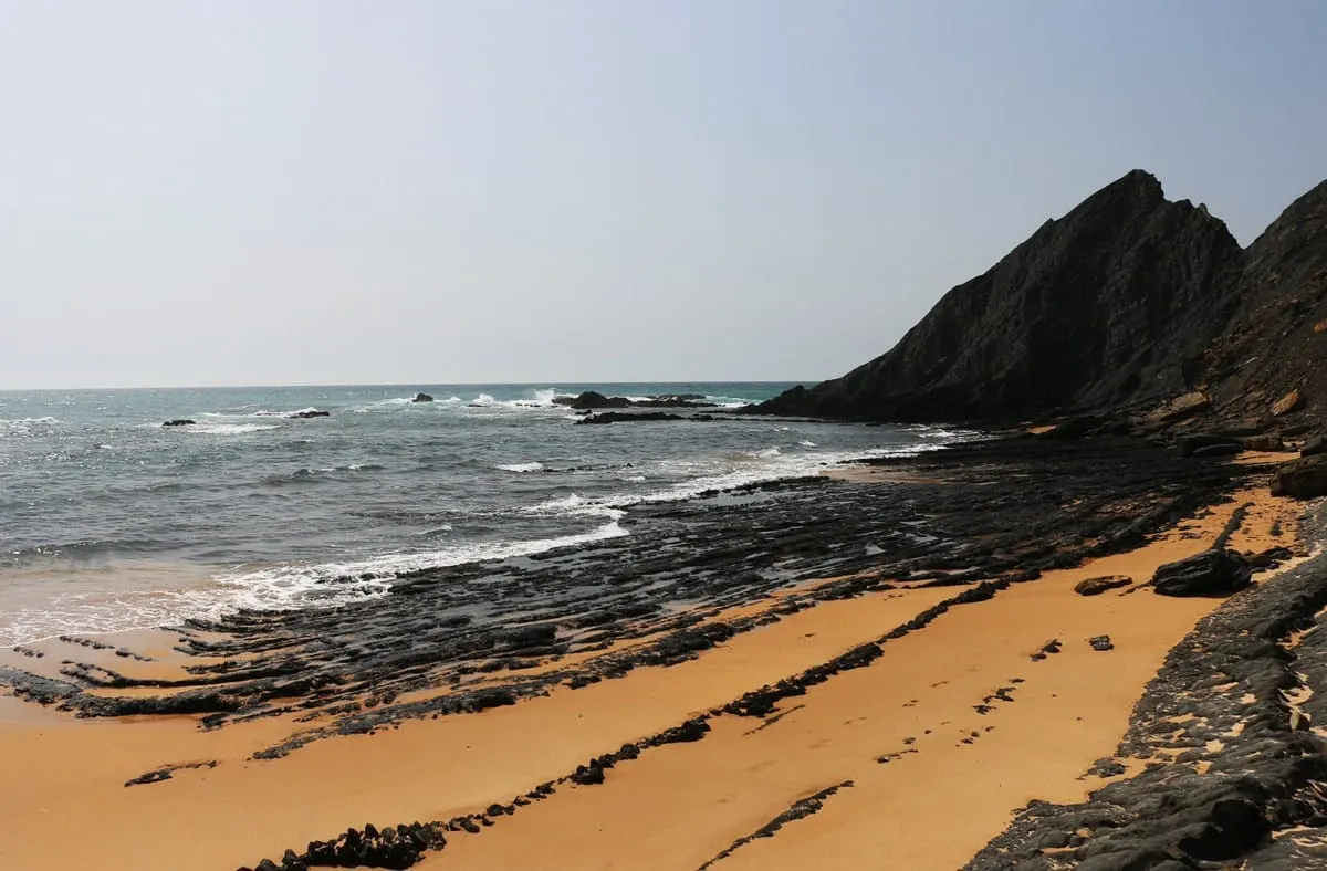 Golden sand beach striped with ridges of black rock beneath a dark headland