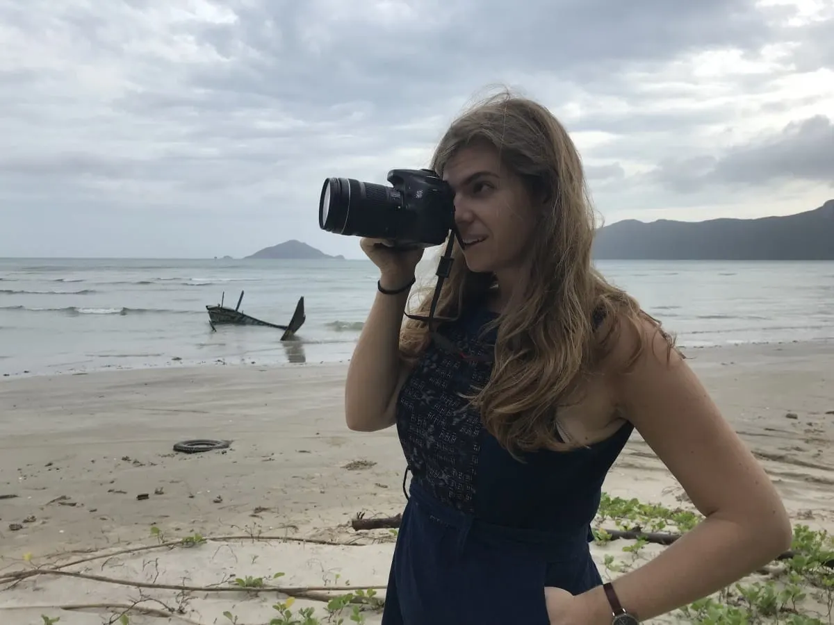 Woman photographing with a DSLR on a beach, a wrecked boat in the surf behind